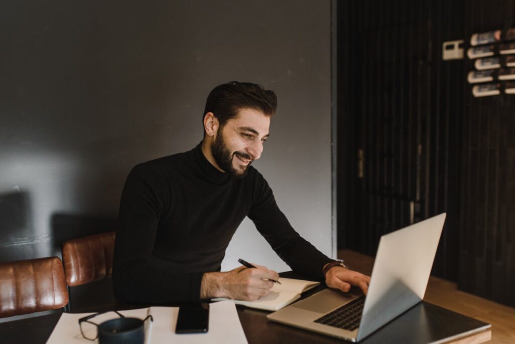 A man with a beard smiling while working on a laptop in a contemporary office setting.